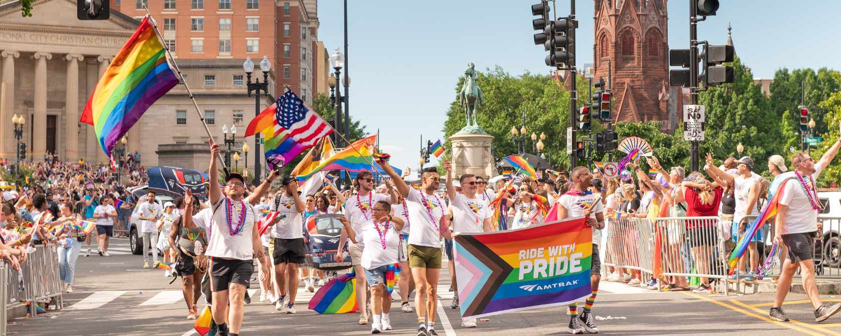 A lively Pride parade with participants in white shirts and colorful beads waving rainbow flags, marching past historic buildings while holding a "Ride with Pride" Amtrak banner.